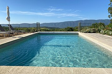 A serene blue swimming pool overlooks distant mountains under a clear sunny sky. Loungers and an umbrella are by the poolside.
