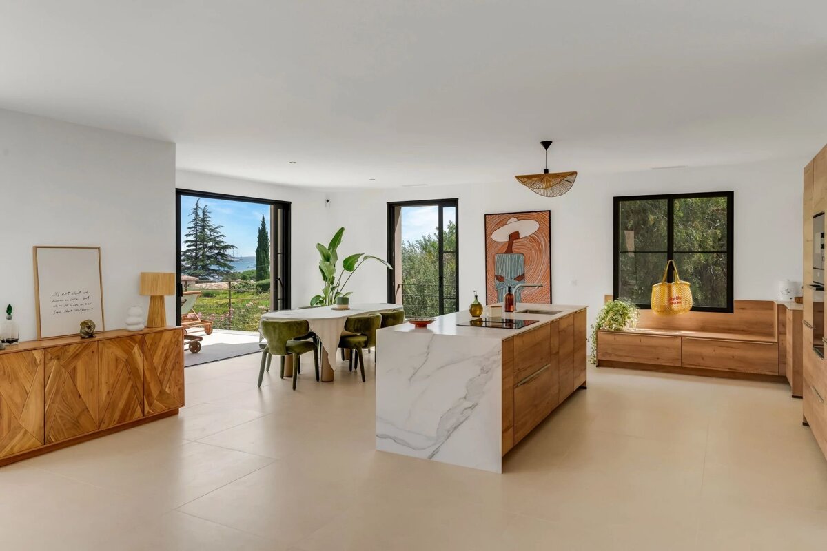 A bright, modern open-plan kitchen and dining room featuring wooden cabinetry, a marble island, and glass doors looking out to a garden.