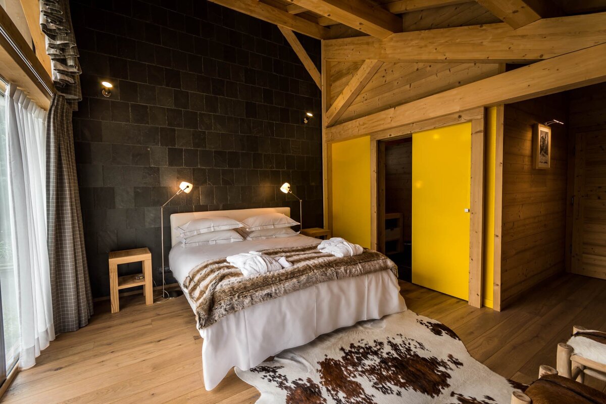 A modern bedroom with dark slate, warm wood, and exposed beams. Features a large bed with faux fur, a cowhide rug, and bright yellow sliding doors.