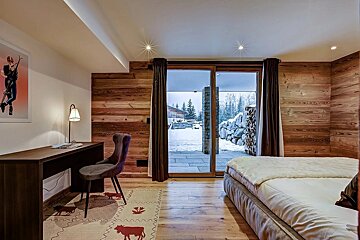 A warm, wood-paneled bedroom with a desk, bed, and a large sliding glass door revealing a snowy winter landscape and pine trees.