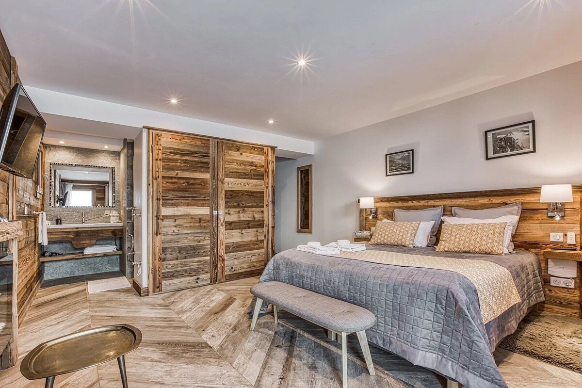 A chic rustic bedroom with wood paneling, herringbone floor, a large bed with gray bedding, and an open-concept bathroom vanity area.