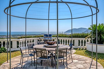 An outdoor stone patio with a dining table under a dome gazebo frame, offering scenic views of the ocean.