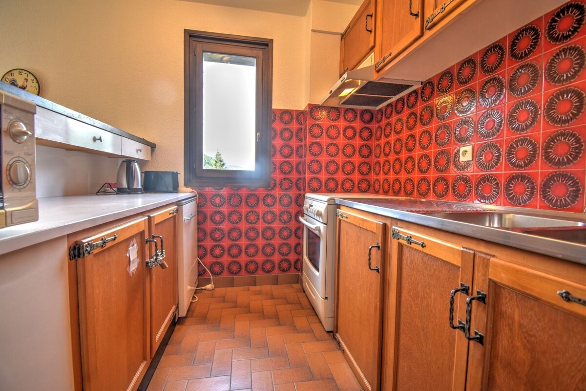 A kitchen with bold red and black patterned wall tiles, wooden cabinets, and a terracotta herringbone floor. A window illuminates the retro-style space.