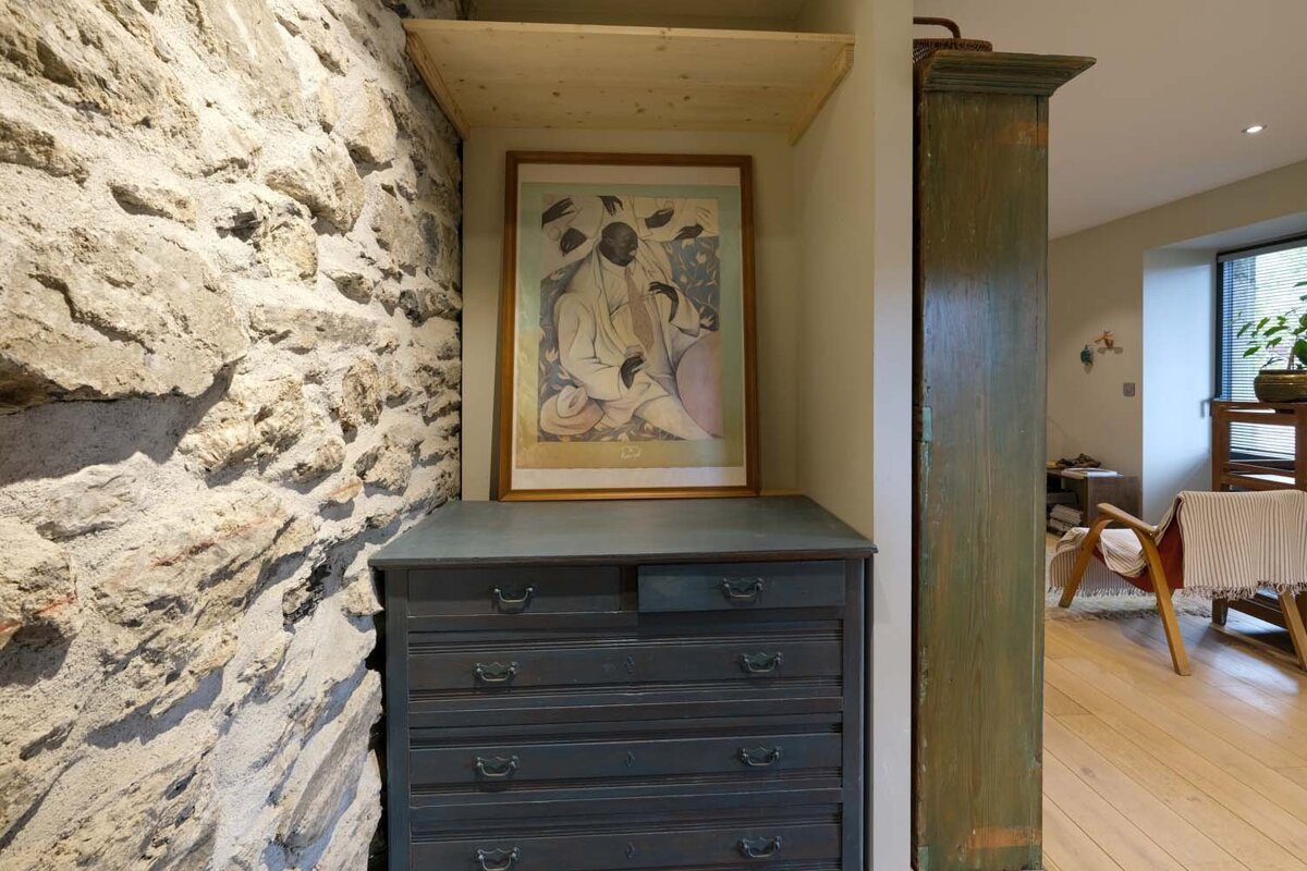 An interior shot featuring a rough stone wall, a dark dresser with a framed artwork and a light wooden shelf above it. A cozy living area is visible to the right.