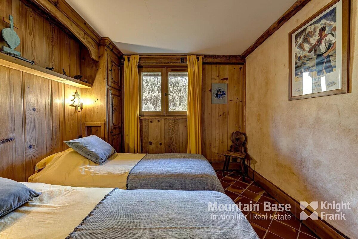 A cozy, rustic twin bedroom with wood-paneled walls, yellow curtains framing a window, and mountain-themed decor. Two beds with yellow sheets.