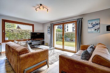 A bright, modern living room featuring two brown sofas, a large TV, and big windows/sliding doors leading to a green garden.