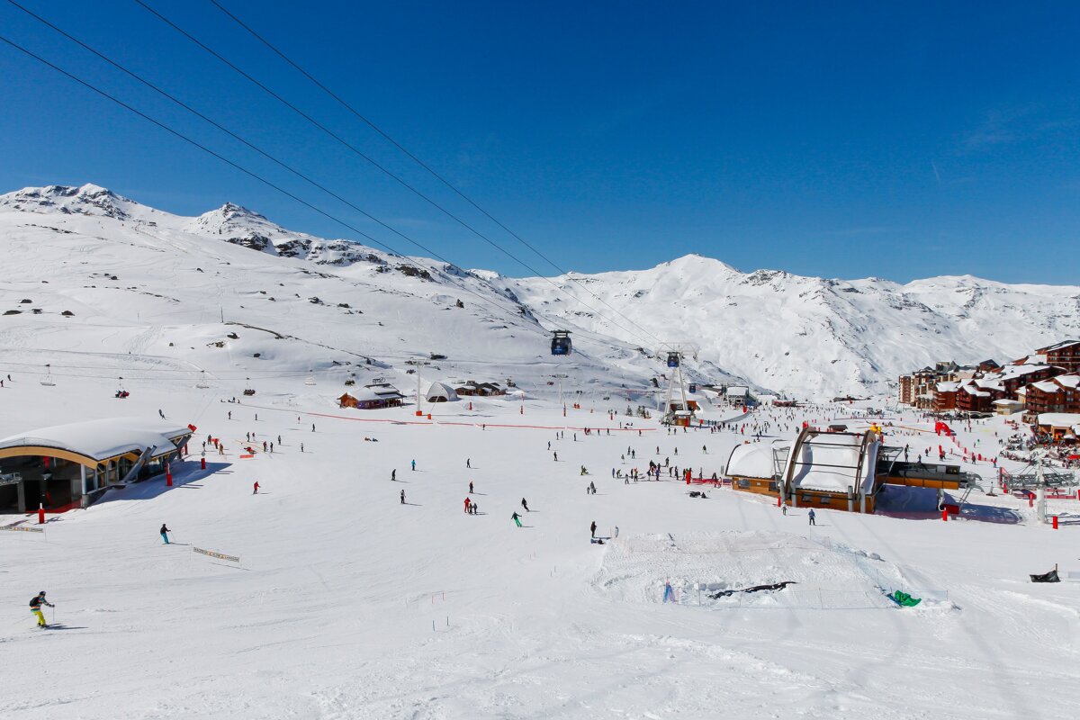 A vast, sunny ski resort nestled among snow-covered mountains, bustling with numerous skiers on slopes, buildings, and gondolas under a clear blue sky.