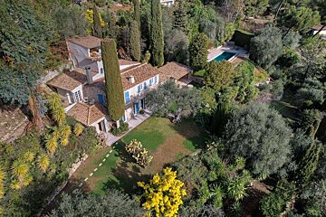 Aerial view of a charming stone house with blue shutters, nestled in a lush, green landscape with tall trees, a sprawling garden, and a swimming pool.
