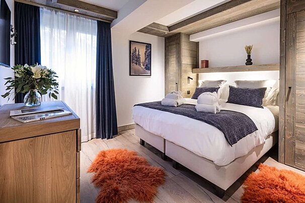 A warm, modern bedroom with a large bed, white linens, blue accents, natural wood paneling, and two fluffy orange rugs on light wood flooring.