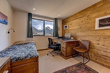 Alpine bedroom with a bed, wooden desk, and a stunning mountain view. Features a wood-paneled wall and cozy decor.