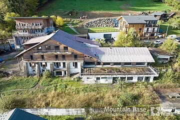 Aerial view of a large, aging wooden mountain building with a long extension, surrounded by greener hills and other structures, including some construction.