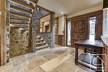 A grand, rustic entryway featuring stone walls and floor, a wooden staircase with illuminated steps, and rich wooden doors and beams.