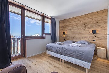 Cozy bedroom featuring a wood-paneled accent wall, a grey bed, and large windows showcasing a stunning mountain view.