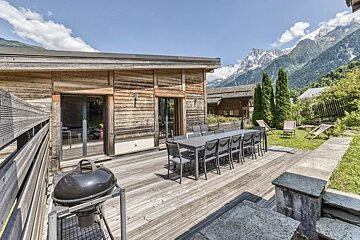 Wooden chalet deck with a dining table and barbecue grill, overlooking lush lawns and snow-capped mountains under a clear sky.