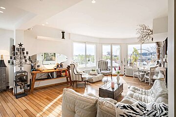 A bright, eclectic living room with white walls, wooden floors, and large windows offering a city view. Features a white sofa and unique furniture.