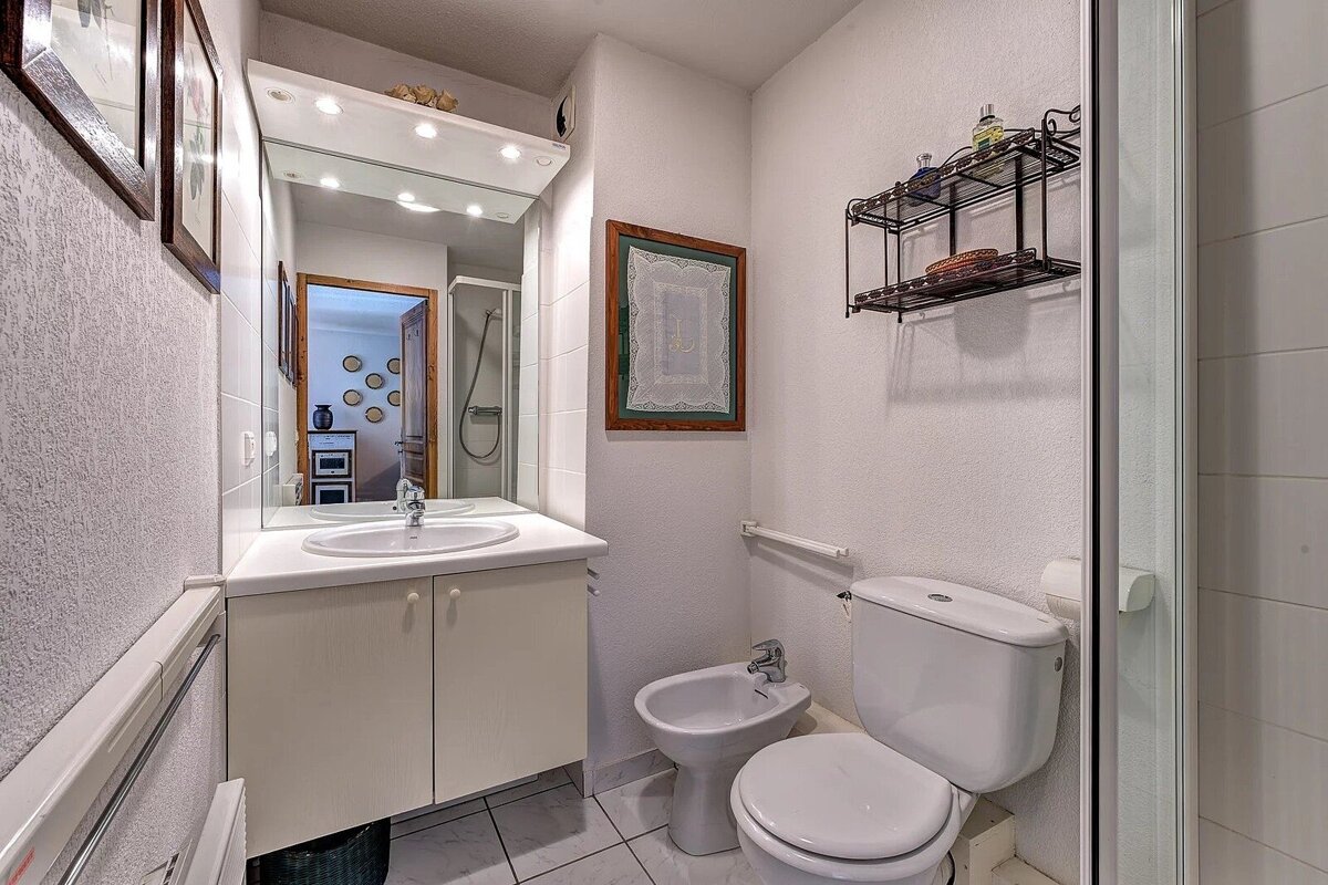 A small, neat bathroom with a white vanity sink, toilet, bidet, lighted mirror, and framed wall art.