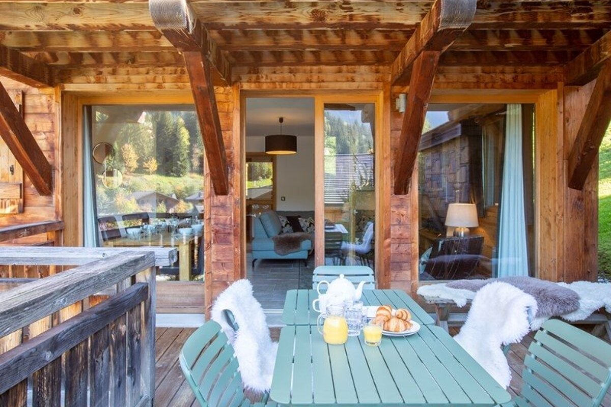 Cozy breakfast setup on a wooden cabin balcony, featuring green chairs with white fur throws, juice, and croissants.