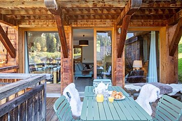 Cozy breakfast setup on a wooden cabin balcony, featuring green chairs with white fur throws, juice, and croissants.