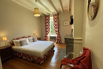 A charming bedroom with a large bed, red patterned curtains, and wooden floors. Features include exposed ceiling beams, a marble fireplace, and elegant decor.