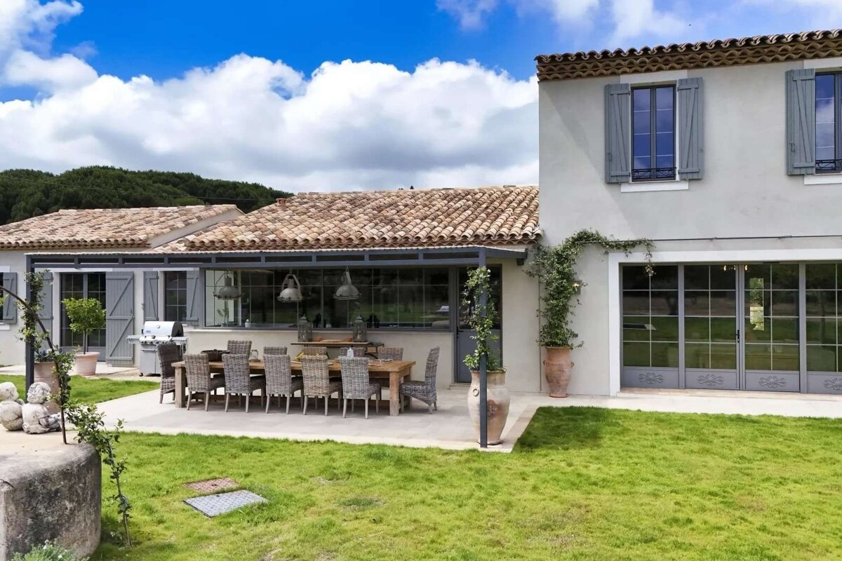 A charming country house with a tiled roof, blue shutters, a large outdoor dining patio, and a vibrant green lawn under a bright, cloudy sky.