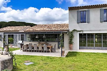 A charming country house with a tiled roof, blue shutters, a large outdoor dining patio, and a vibrant green lawn under a bright, cloudy sky.
