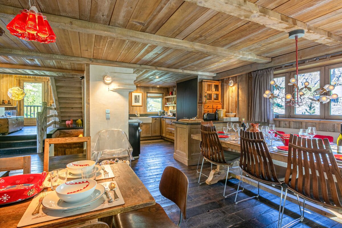 Rustic wooden interior with dining and kitchen areas, featuring unique lighting, set tables, and a view to nature.