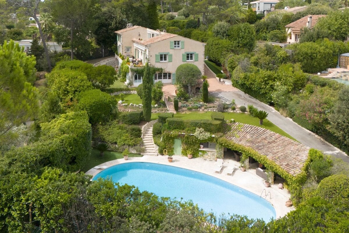 Aerial view of a luxury villa with a curved swimming pool, lush terraced gardens, and a stone house with light green shutters.