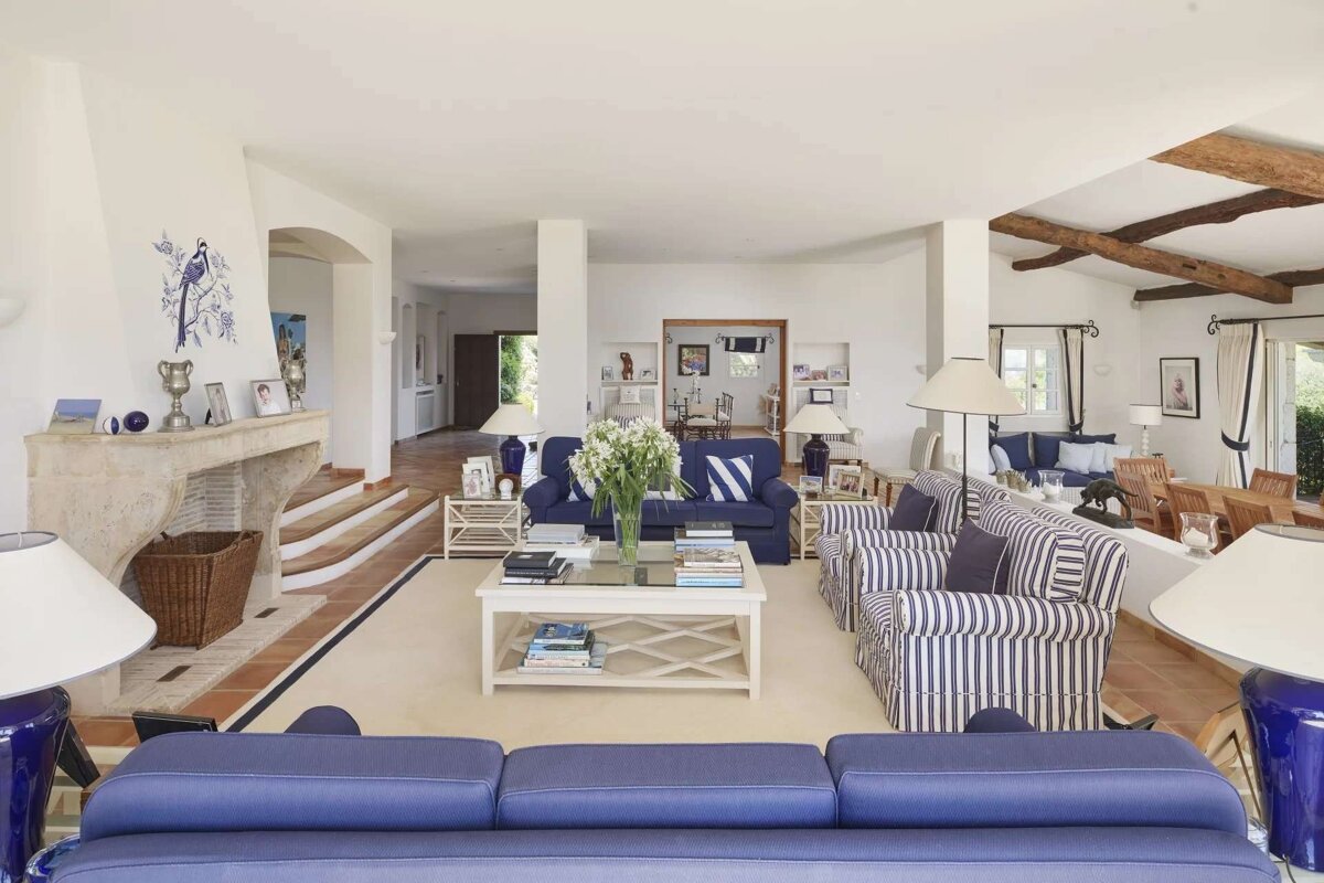 A spacious, bright living room with a blue and white theme, featuring blue sofas, striped armchairs, a fireplace with bird art, and exposed wooden beams.