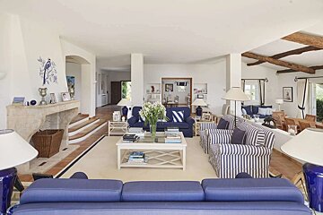 A spacious, bright living room with a blue and white theme, featuring blue sofas, striped armchairs, a fireplace with bird art, and exposed wooden beams.
