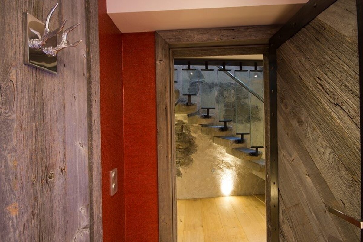 Rustic wooden door opens to a modern staircase with floating steps, glass railings, and stone walls illuminated from below.