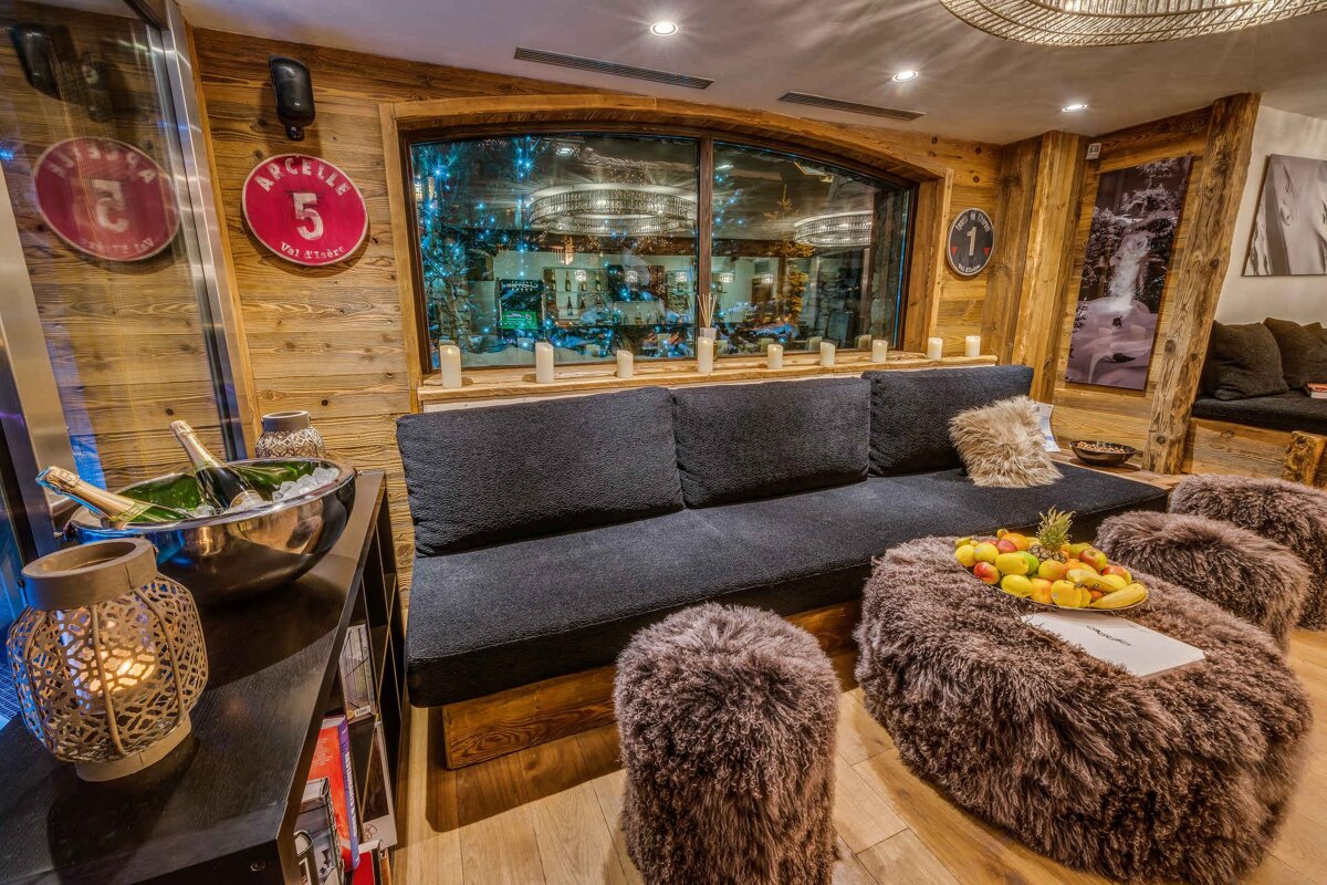 Cozy rustic lounge with wood-paneled walls, a large black sofa, furry ottomans, a fruit platter, and champagne chilling in an ice bucket.