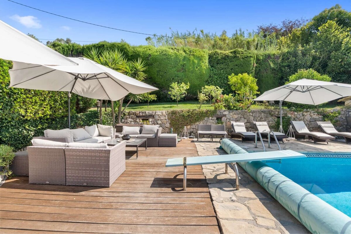 A sunny poolside patio with a blue swimming pool, diving board, and wooden deck. Comfortable wicker sofas under umbrellas, lounge chairs, and lush green hedges complete the scene.