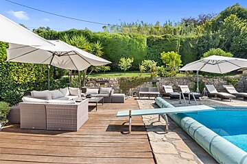 A sunny poolside patio with a blue swimming pool, diving board, and wooden deck. Comfortable wicker sofas under umbrellas, lounge chairs, and lush green hedges complete the scene.