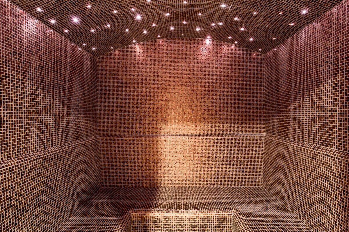 A warm brown mosaic-tiled steam room with an arched ceiling sparkling with star-like lights. Built-in benches line the walls.