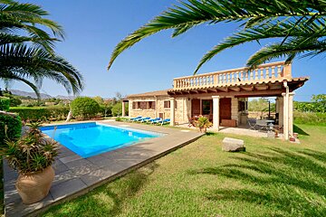 A sunny villa with a bright blue pool, green lawn, and palm trees under a clear sky, with distant mountains in the background.