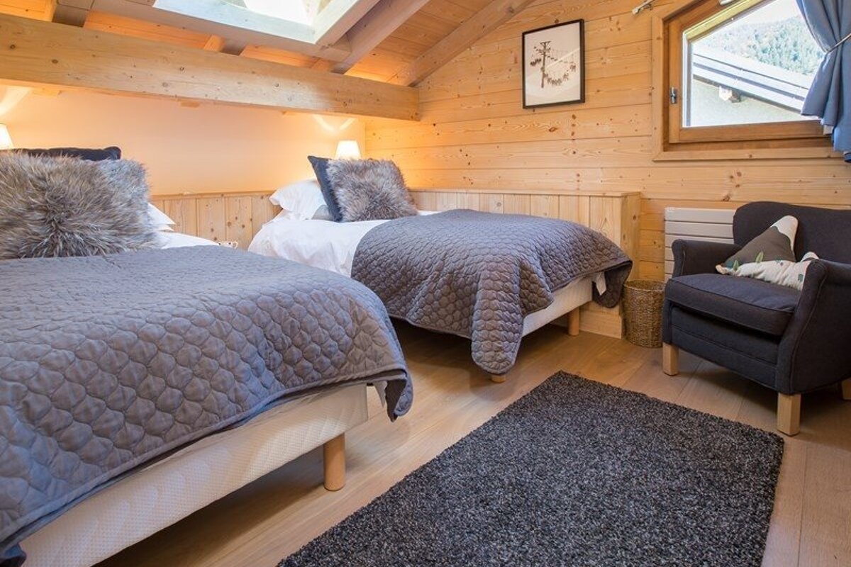 Cozy cabin bedroom with wooden walls, two twin beds with gray quilted blankets and fuzzy pillows, and a dark armchair.