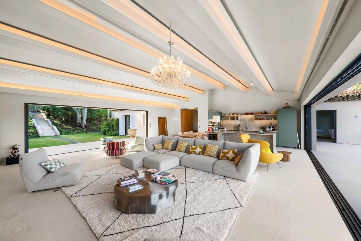 A spacious, modern open-plan living room with a large sofa, chandelier, kitchen, and a view of a lush garden through wide glass doors.