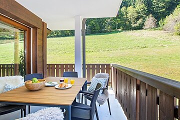 A cozy wooden balcony with a set breakfast table overlooking a lush green field and forest.
