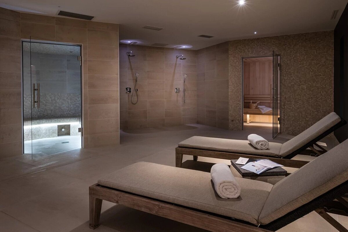 A modern spa relaxation area featuring two lounge chairs, a glass-door sauna, a steam room, and multiple showers in calming neutral tones.