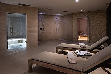 A modern, tiled spa area featuring two loungers, multiple showers, a brightly lit steam room, and a cozy sauna, all in warm, earthy tones.
