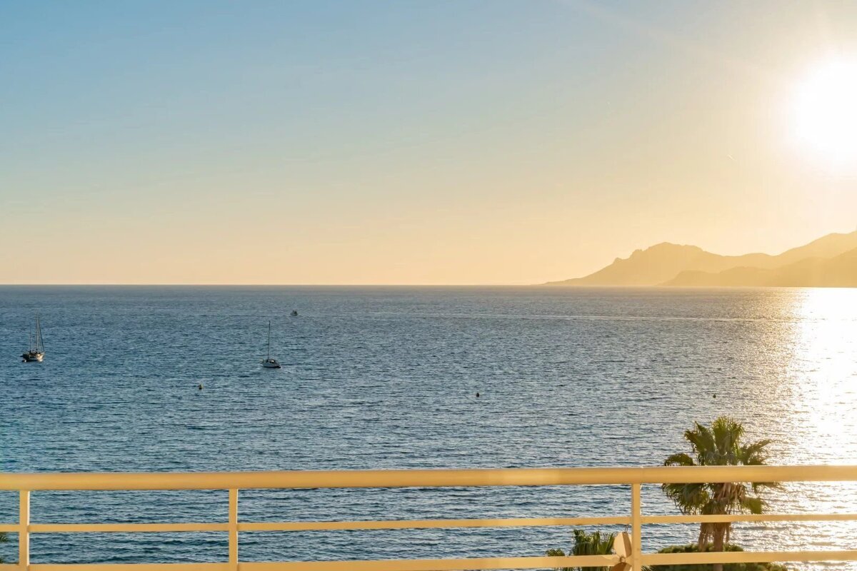 Ocean view from a balcony with sailboats, a palm tree, and distant mountains under a warm, glowing sun.