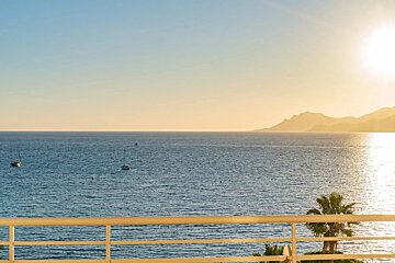 Ocean view from a balcony with sailboats, a palm tree, and distant mountains under a warm, glowing sun.