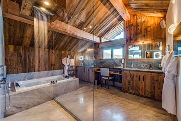 Luxury wooden bathroom with a glass shower, large tub, sloped ceiling, dual vanities, and natural light.