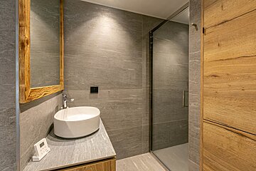 A modern bathroom with grey tiled walls, a white vessel sink, a wooden-framed mirror, and a glass-enclosed shower.