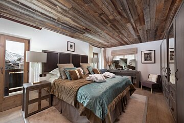 A luxurious bedroom featuring a rustic wood-paneled ceiling, a large bed, elegant decor, and a balcony with views of snow-capped mountains.