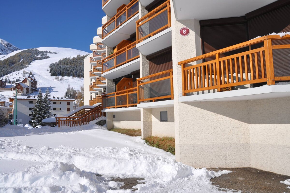 A modern apartment building with wooden balconies in a snowy mountain resort. Bright sunshine illuminates the snow-covered ground and distant ski slopes.
