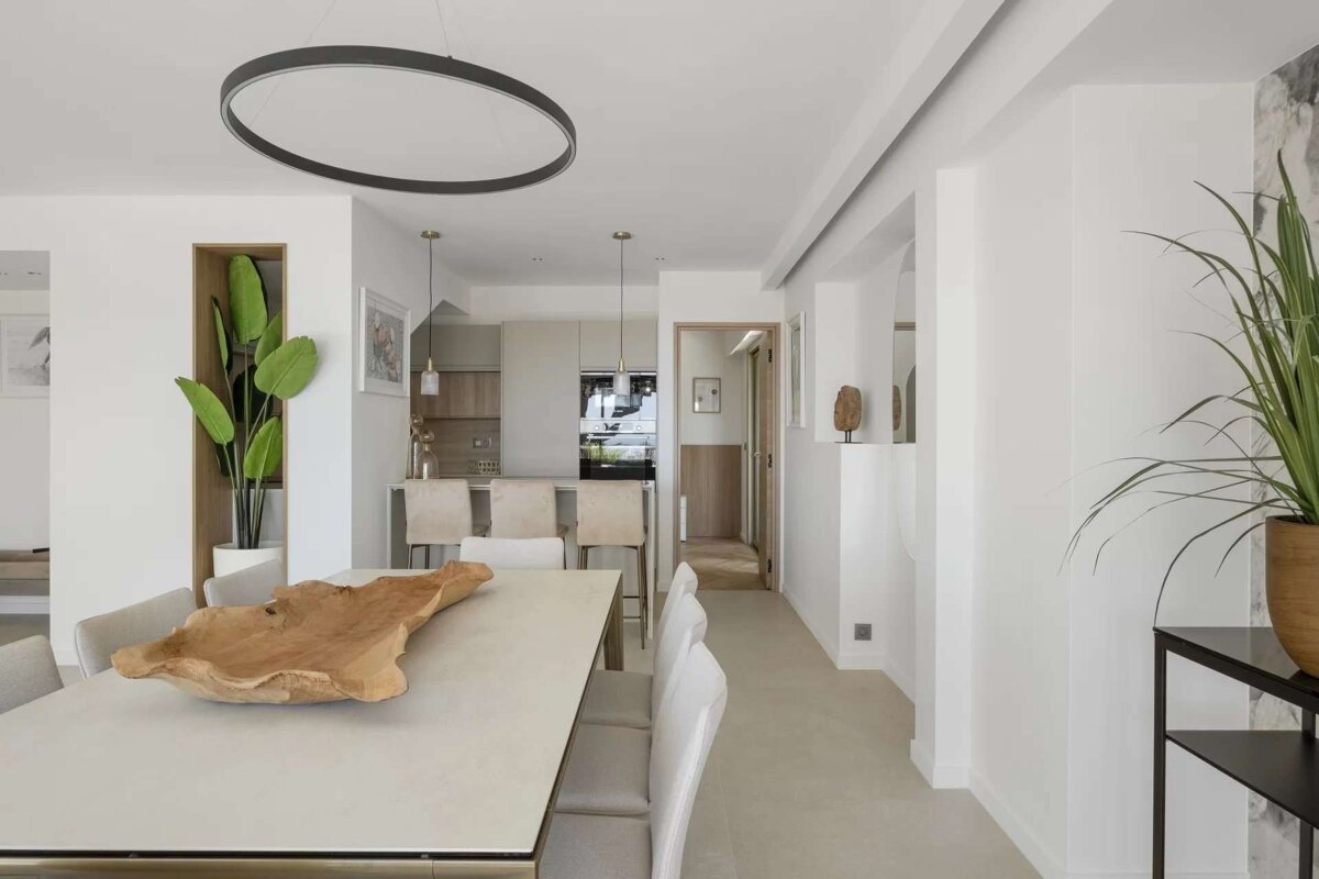 Stylish open-plan dining area and kitchen with white walls, light furniture, green plants, and a black oval chandelier.