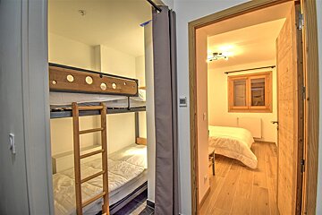 A cozy interior features bunk beds partially hidden by a curtain on the left, and a bedroom with a double bed, wooden window, and light wood floor on the right.