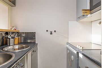 A compact kitchen with dark counters, two stainless steel sinks, and visible plumbing on a white wall. Utensils and a dishwasher are also seen.