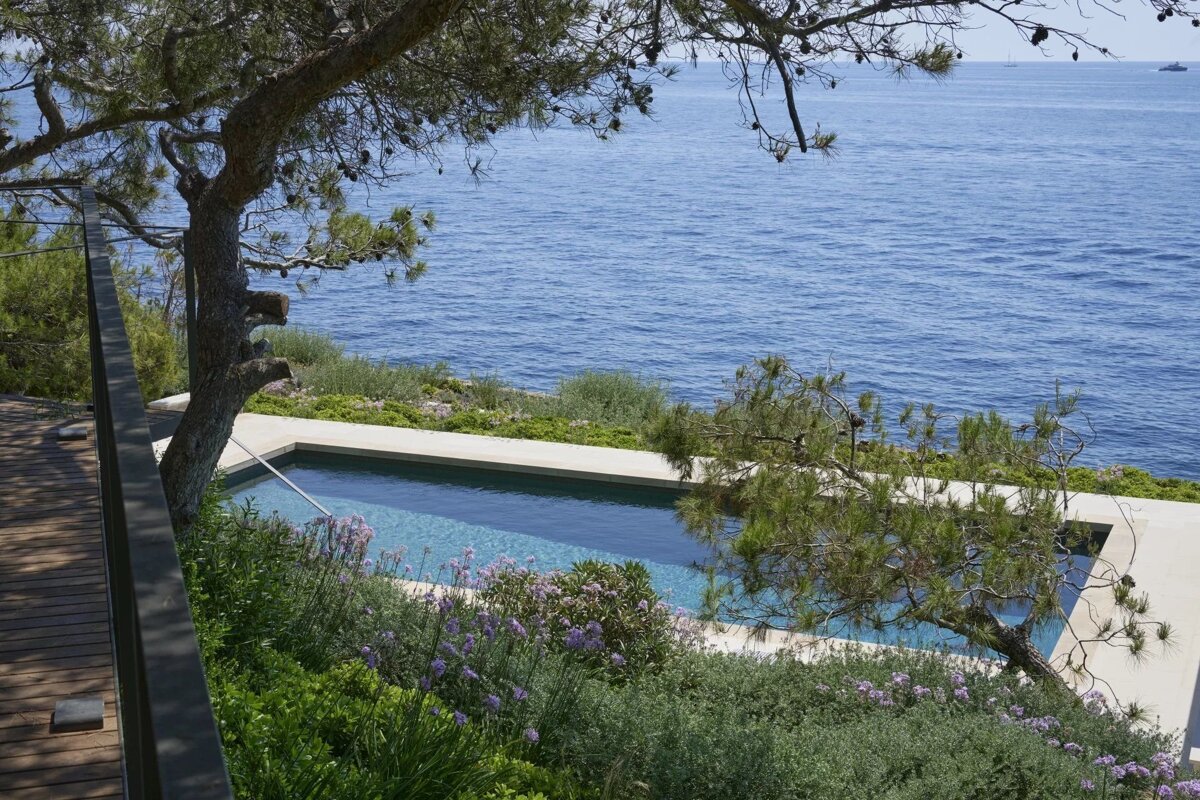 A high-angle view of a swimming pool situated on a cliffside overlooking the blue sea, surrounded by trees and greenery.